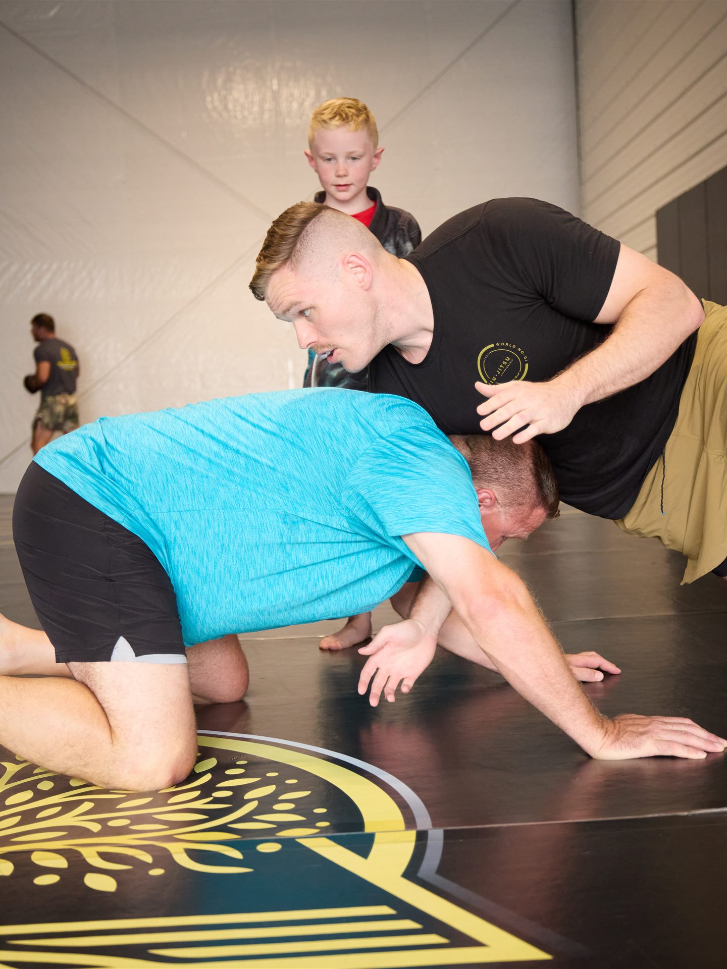 A young wrestler watching closely as a coach demonstrates a takedown on the Golden Oak logo mat